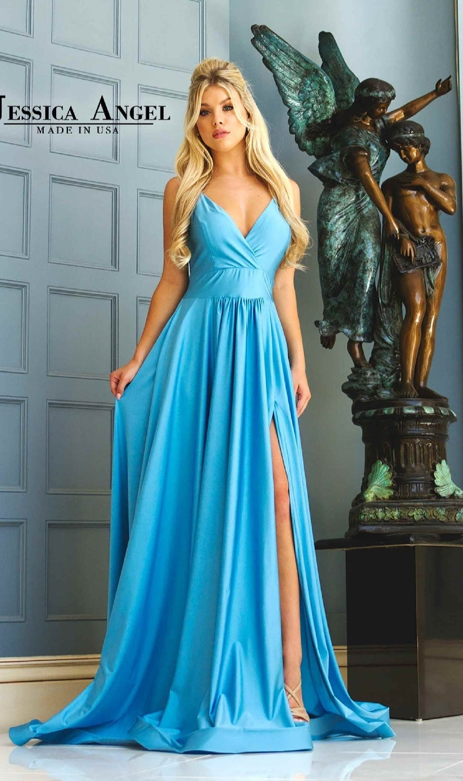 A woman in a long, light blue V-Neck A-Line Prom Dress (Jessica Angel 343) with a thigh-high slit stands indoors beside a bronze statue of two angels, against paneled walls bearing the